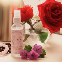 Picture shares a picture of the Emisha Wellness Rose Water in a 60ml size. This particular one is set with the backdrop of life with two beautiful & vibrant red roses next to Tiger's Eye crystals resting close by. The Rose and Geranium Water is also restfully present while gently announcing its soft presence with tiger's eye and rose quartz to add some loving and passionate energy to the conversation.